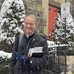 Fr De Lion stands on a snowy day outside the Church. The red door entrance is behind his left shoulder.